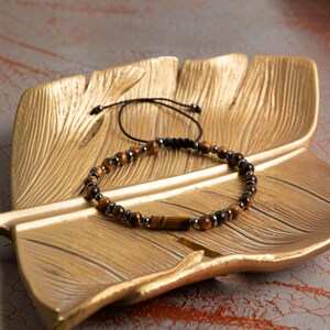 May include: A beaded bracelet featuring alternating round brown tiger eye beads and small metallic gray beads, with a single cylindrical tiger eye bead at the center. The bracelet includes a black cord with an adjustable sliding knot closure.
