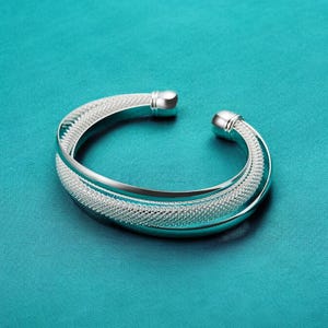 May include: A silver bangle bracelet with a woven design. The bracelet is open and has a smooth, polished finish.