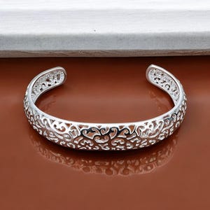 Pode incluir: Um bracelete de prata com um design floral delicado e intrincado. O bracelete é aberto e tem um estilo simples e elegante.
