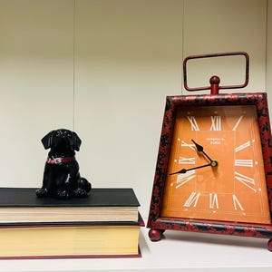 May include: A black ceramic dog figurine with a red collar sits atop two books. A red and black metal clock with Roman numerals and the text "Antique de Paris" is also present. The clock has a rectangular face and a handle.