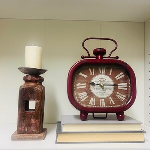 May include: A red metal alarm clock with a white face and black Roman numeral hour markers. The clock has a crown and the text "The Clock on the Wall, London 1861" on the face. The clock is sitting on a stack of books on a white shelf.
