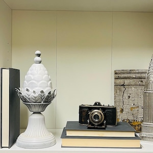 May include: A white decorative pineapple-shaped object with a pedestal base, a vintage camera, a silver model of the Chrysler Building, and two books are displayed on a shelf. The objects are arranged against a neutral background.