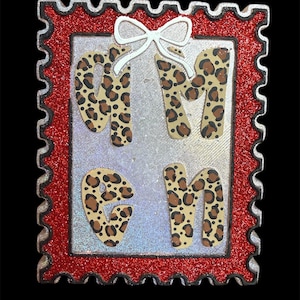 May include: A rectangular decorative piece with a red glitter border and a silver interior. The letters "GMGN" are in the center, with a leopard print pattern. A white bow is at the top.