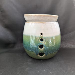 May include: A ceramic oil warmer with a white top, a white middle section, and a green base. The warmer has three circular holes in the front. The top is a separate piece, and the base is a rounded shape.