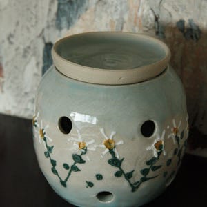 May include: A pale blue ceramic oil warmer with a lid. The round warmer has a daisy design with white petals and yellow centres, and green stems. It has several holes for ventilation.