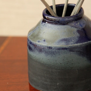 May include: A handmade ceramic vase with a dark blue and grey gradient glaze. The vase has a wide base and a narrow opening, with several light-coloured reeds or sticks inserted. The vase sits on a reddish-brown surface.