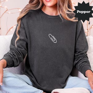 May include: Dark gray long-sleeve shirt with a white paperclip graphic on the chest. The shirt has a relaxed fit and a vintage wash. The word "Pepper" is in a black starburst shape.