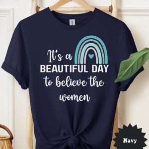 May include: Navy blue t-shirt with the text "It's a BEAUTIFUL DAY to believe the women" and a turquoise rainbow graphic. The shirt is knotted at the waist and hangs on a wooden hanger.