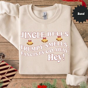 May include: Sand-colored sweatshirt with the text "Jingle Bells, Trumpy Smells, Fascists Go Away, Hey!" in red and white. The design includes three gold bells with red bows. The sweatshirt is folded on a wooden surface.