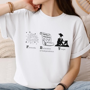May include: White t-shirt with a black graphic design featuring fireworks, a scroll with the text "Declaration of Independence", and a group of soldiers labeled "Troops".