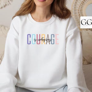 May include: White sweatshirt with the text "COURAGE is contagious" in rainbow colors.