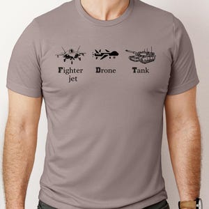 May include: A heather grey t-shirt with a black graphic of a fighter jet, drone, and tank. The text "Fighter jet", "Drone", and "Tank" is printed below each graphic.
