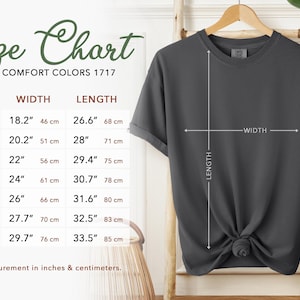 Puede incluir: Una camiseta gris oscuro cuelga de un perchero de madera, con una tabla de tallas a la izquierda. La tabla enumera las tallas S a 4XL, con medidas en pulgadas y cent&iacute;metros. Se indica el ancho y el largo de la camiseta.