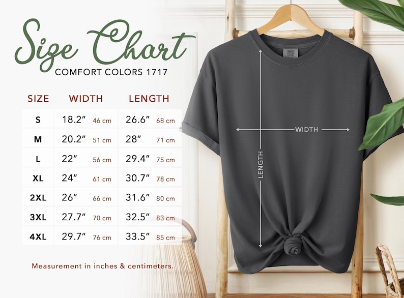 Puede incluir: Una camiseta gris oscuro cuelga de un perchero de madera, con una tabla de tallas a la izquierda. La tabla enumera las tallas S a 4XL, con medidas en pulgadas y cent&iacute;metros. Se indica el ancho y el largo de la camiseta.