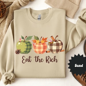 May include: Sand-colored sweatshirt featuring three apple-shaped designs with plaid patterns, leaves, and worms. The text "Eat the Rich" is printed below the apples. A pinecone and cinnamon sticks are also visible.