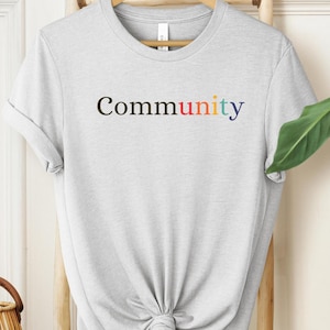 May include: A gray t-shirt with the word "community" printed on it in black letters with rainbow colored accents.