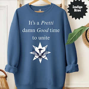 May include: Indigo blue sweatshirt with the text "It's a Pretti damn Good time to unite" in white. The design includes a star and bird graphic. The sweatshirt is displayed on a wooden rack.