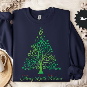 May include: Navy blue sweatshirt featuring a green and yellow Christmas tree design with stars and swirls. The text "Merry Little Solstice" is printed at the bottom of the tree. The sweatshirt is a crew neck style.