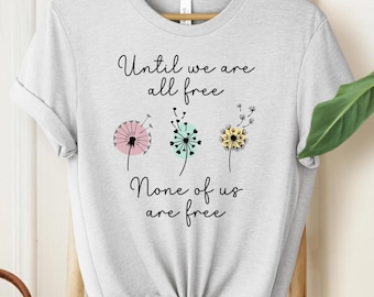 May include: A light gray t-shirt with the text "Until we are all free None of us are free" printed on it. The text is in black and there are three stylized dandelion illustrations in pink, green, and yellow.