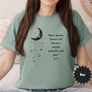 May include: Sage green t-shirt featuring a black crescent moon and star design with the text "How many hours of sleep a night should you get? 6-7". The shirt has rolled-up sleeves and a relaxed fit.