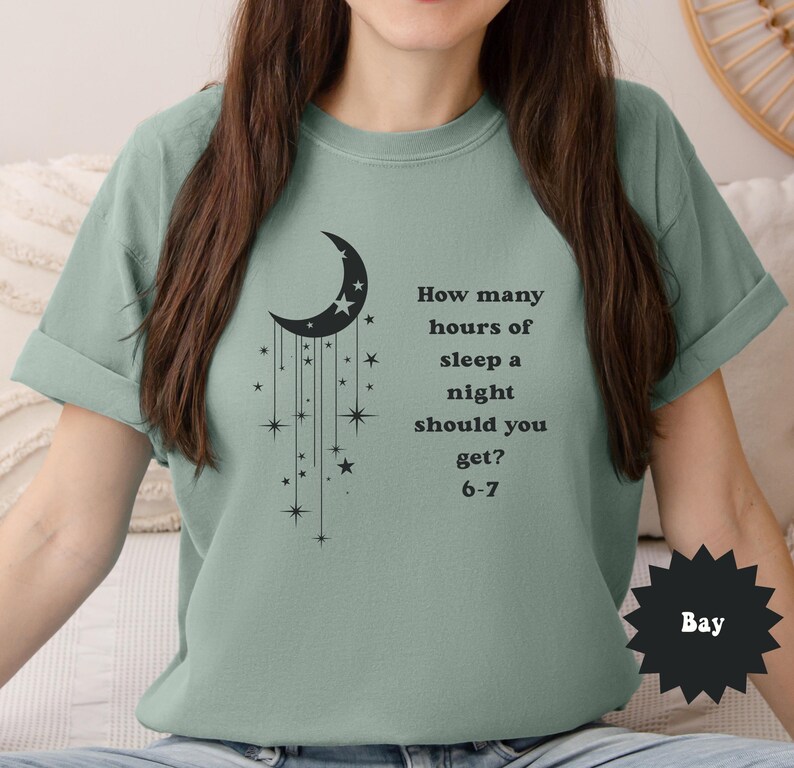 May include: Sage green t-shirt featuring a black crescent moon and star design with the text "How many hours of sleep a night should you get? 6-7". The shirt has rolled-up sleeves and a relaxed fit.
