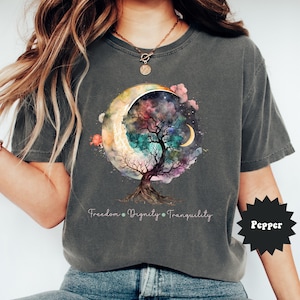 May include: Dark gray t-shirt featuring a colorful tree design within a crescent moon, with the words "Freedom, Dignity, Tranquility" below. The shirt has a relaxed fit and short sleeves. The word "Pepper" is printed on a black starburst.