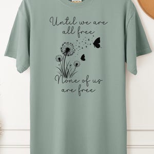 May include: Sage green t-shirt with a black graphic of dandelions and butterflies. The text on the shirt reads "Until we are all free. None of us are free."