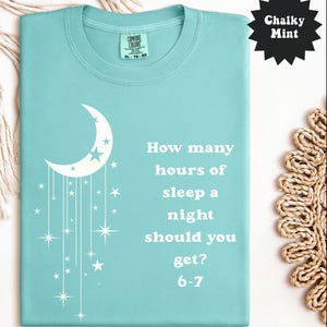 May include: A light teal Comfort Colors t-shirt with a white crescent moon and stars design. The shirt features the text "How many hours of sleep a night should you get? 6-7". The shirt is a size XL-TG-EG.