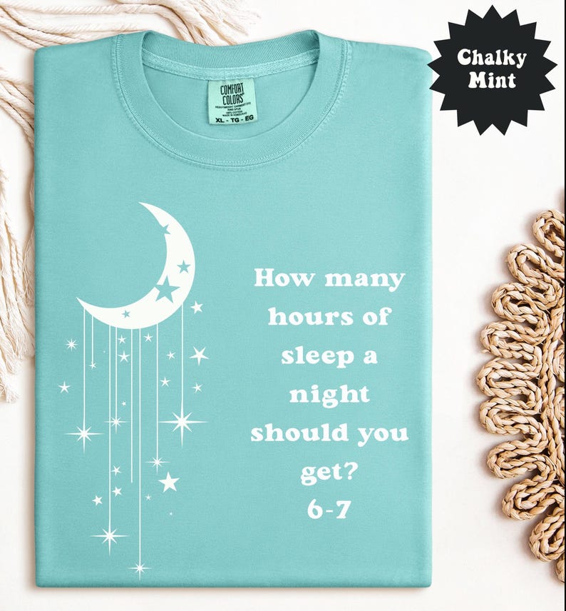 May include: A light teal Comfort Colors t-shirt with a white crescent moon and stars design. The shirt features the text "How many hours of sleep a night should you get? 6-7". The shirt is a size XL-TG-EG.