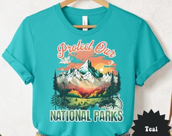 May include: Teal t-shirt with a graphic of a mountain landscape, trees, and the text "Protect Our National Parks." The design features a sunset and a compass. The shirt is displayed on a wooden hanger.