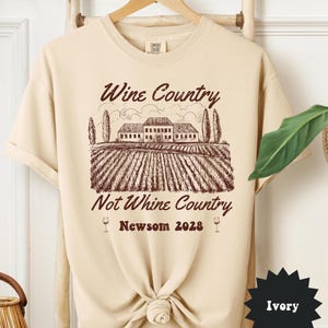 May include: Ivory-colored Comfort Colors t-shirt with a brown graphic. The graphic reads "Wine Country" above a vineyard and building illustration, with "Not Whine Country" and "Newsom 2028" below. The shirt is hanging on a wooden rack.