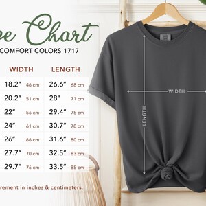 May include: A dark gray t-shirt displayed on a wooden hanger, with a size chart to the left. The size chart shows measurements in inches and centimeters for width and length, ranging from size S to 4XL.