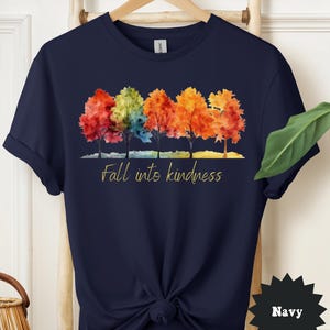 May include: Navy blue t-shirt featuring a watercolor design of five trees with colorful autumn foliage. The trees are in shades of red, orange, yellow, and green. The text "Fall into kindness" is printed in gold script below the trees.