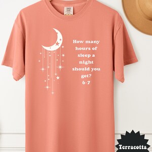 May include: Terracotta-colored t-shirt with a white crescent moon and star design. The shirt features the text: "How many hours of sleep a night should you get? 6-7". The shirt is displayed on a wooden hanger.