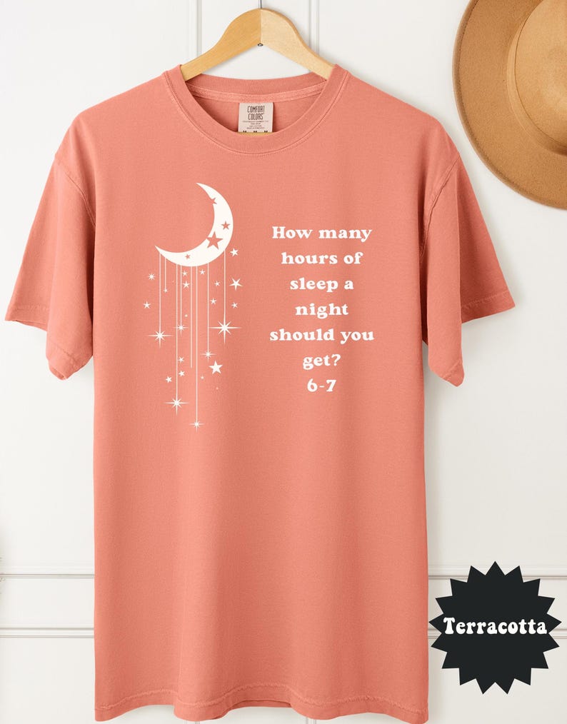 May include: Terracotta-colored t-shirt with a white crescent moon and star design. The shirt features the text: "How many hours of sleep a night should you get? 6-7". The shirt is displayed on a wooden hanger.