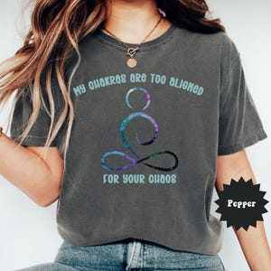 May include: Dark grey t-shirt with the text "My chakras are too aligned for your chaos" in light blue. The shirt features a colorful, cosmic-themed yoga pose graphic. The word "Pepper" is in a black starburst.