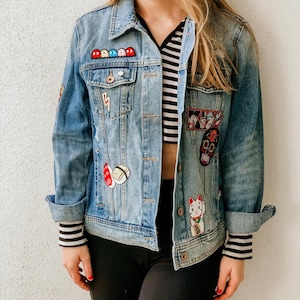 May include: Light blue denim jacket with various embroidered patches, including a lightning bolt, a skull, and a lucky cat. The jacket has a classic collar, button-up front, and rolled-up sleeves with striped cuffs.