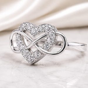 May include: A silver-toned ring featuring a central heart-shaped motif integrated with an infinity symbol. The heart is embellished with a series of small, clear, round-cut stones set along its perimeter, while the band remains smooth and polished.