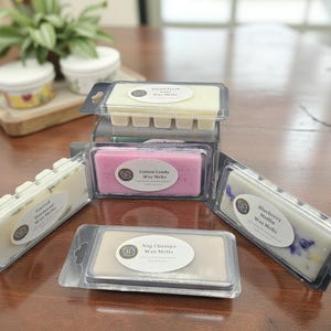 May include: An assortment of wax melts in clear plastic packaging. The scents include Island Fresh, Cotton Candy, Nag Champa, and Blueberry Muffin. Each package has a label with the scent name. The wax melts are arranged on a wooden table.