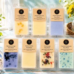 May include: Seven rectangular clear plastic clamshell containers of wax melts arranged in two rows. Each features a tan label with the SS logo, scent name, and Net Wt. 3.2 - 3.4 oz. Scents include Crème Brulee, Island Fresh Gain, Fabric Softener, Blackberry Sage, Honey Vanilla, Froot Loops, and Tropical Daydream.