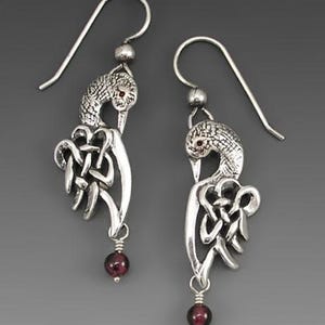 May include: A pair of silver drop earrings featuring a bird motif with intricate knotwork detailing on the wings. Each earring is accented with a small, round, deep red garnet bead suspended at the bottom and a matching red stone eye.