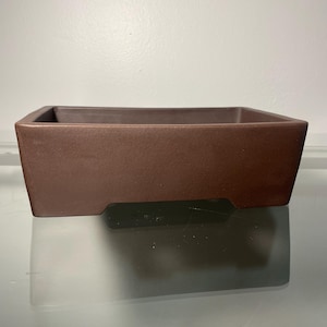 May include: A rectangular brown ceramic bonsai pot with a slightly raised lip around the top edge. The pot has a matte finish and a slightly textured surface.