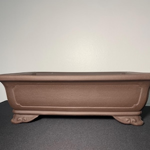 May include: A rectangular brown ceramic bonsai pot with a textured surface and decorative feet.
