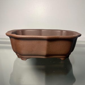 May include: A brown ceramic bonsai pot with a rounded rectangular shape and a slightly flared rim. The pot has eight sides and rests on three small feet.