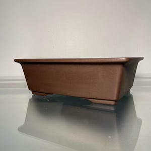 May include: A rectangular brown bonsai pot with a slightly flared rim. The pot is made of a matte material and sits on a reflective surface. The pot is designed for growing small trees and plants.