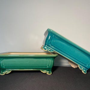 May include: Two rectangular ceramic bonsai pots. One is upright, the other is tilted. Both are teal green with a beige interior and decorative feet. The pots have a glossy finish and are suitable for growing small plants.