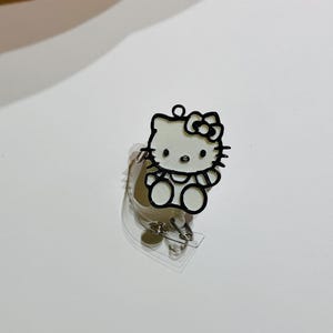 May include: A white Hello Kitty character with black outlines and a red bow is attached to a clear plastic badge holder.