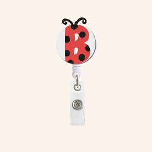 Könnte beinhalten: Ein rotes und schwarzes Marienkäfer-Badge-Rolle mit dem Buchstaben "B" auf weißem Hintergrund. Die Rolle hat ein einziehbares Kabel und einen Clip zum Befestigen an Kleidung oder Accessoires. Das Design ist verspielt.