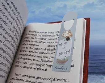 May include: A light blue bookmark with the text "Let go and let God" and a floral and bird nest design. The bookmark is inserted into a book with printed text. The background shows a sky and ocean.