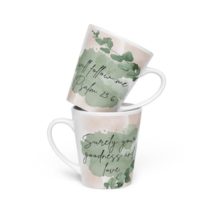 Może przedstawiać: Dwa białe ceramiczne kubki do latte ułożone jeden na drugim, każdy z uchwytem. Jeden kubek ma napis "will follow me Psalm 23:6", a drugi "Surely your goodness and love". Oba kubki mają akwarelowy wzór z zielonymi liśćmi i brzoskwiniowym tłem.